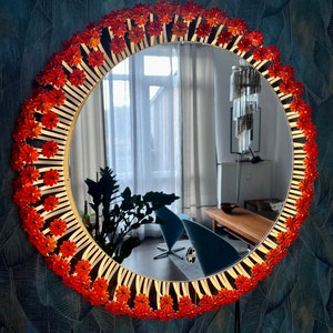 Op de afbeelding: Een ronde spiegel met een gouden lijst versierd met oranje bloemvormige versieringen. De spiegel reflecteert een kamer met een raam en meubels.