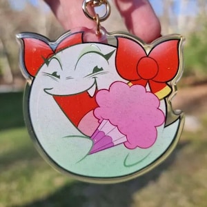 Puede incluir: Un fantasma blanco con lazos rojos y un algodón de azúcar rosa en la mano. El fantasma sonríe con la boca abierta y tiene dientes afilados. El fondo está borroso y es verde.