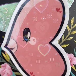 May include: A pink heart-shaped sticker with a white outline and a glittery finish. The sticker has a cartoon face with a blue eye and a pink nose. The background of the sticker is filled with small pink hearts and a pattern of white squares.