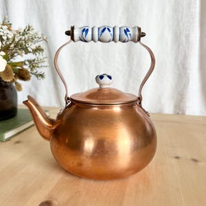 May include: A copper tea kettle with a rounded body and a curved spout. The handle is made of copper and has a white ceramic grip with blue floral designs. The lid is topped with a small, white ceramic knob with blue accents.