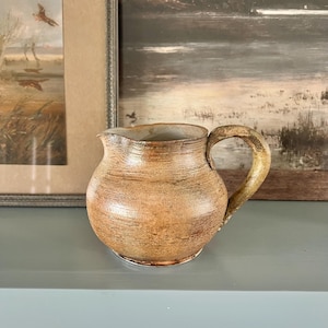 May include: A small, handcrafted ceramic pitcher with a rounded body and a curved handle. The pitcher has a speckled, earthy brown and tan color palette, with visible texture. The spout is slightly flared. A rustic, vintage-style decorative item.