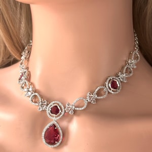 Könnte beinhalten: Eine silberne Halskette mit einem tropfenförmigen Anhänger, der einen großen roten Edelstein enthält, der von kleineren klaren Edelsteinen umgeben ist. Die Halskette hat eine zarte Kette mit kleinen klaren Edelsteinen und filigranen Designs.