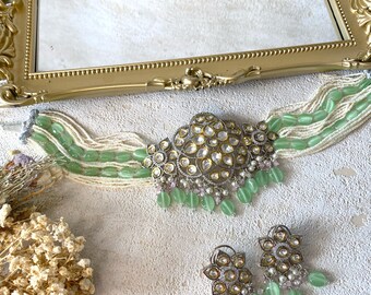 Op de afbeelding: Een zilveren en gouden ketting met groene en witte kralen. De ketting heeft een grote, sierlijke hanger met heldere en witte stenen. De ketting wordt geleverd met bijpassende oorbellen.