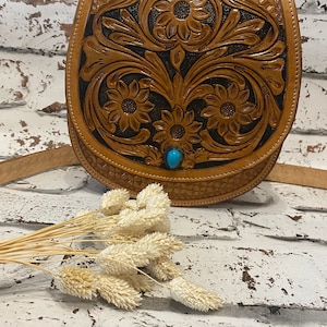 May include: A brown leather purse with a floral design and a turquoise stone accent. The purse has a long strap and is made of tooled leather.