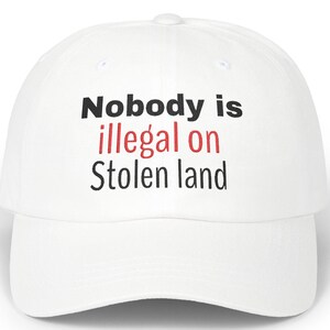 May include: A white baseball cap featuring the text "Nobody is illegal on Stolen land" in black and red. The cap has a curved peak and a button on top. A classic, casual hat.