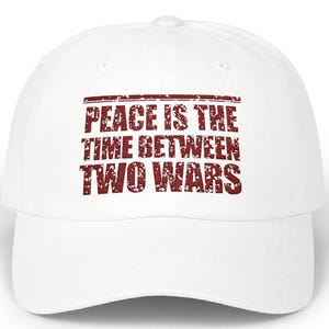 Puede incluir: Gorra de béisbol blanca con el texto "PEACE IS THE TIME BETWEEN TWO WARS" en letras granates desgastadas. La gorra tiene una visera curva y un diseño clásico de seis paneles.