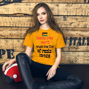 Può includere: Una donna indossa una maglietta gialla con una bandiera nera e verde e il testo "Taking lives won't crush the fire of resistance" stampato su di essa. È seduta su un pavimento di legno con un casco rosso accanto a lei.