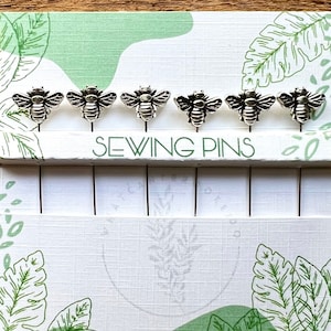 May include: A set of six silver bee-shaped sewing pins in a white and green card packaging with the text "Sewing Pins".