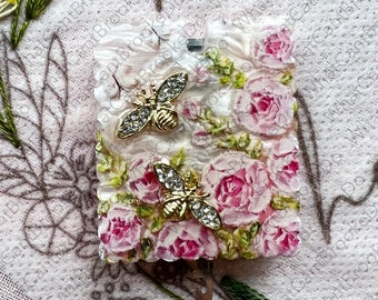 May include: A square, floral-themed object with pink roses and two gold-colored bee embellishments. The object has a textured, possibly resin-like surface. Green embroidery and other floral elements surround the object.