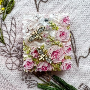 May include: A square, floral-themed object with pink roses and two gold-colored bee embellishments. The object has a textured, possibly resin-like surface. Green embroidery and other floral elements surround the object.