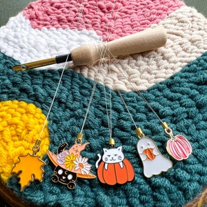 May include: Five colorful enamel pins with different Halloween themes hanging from a tapestry. The pins include a witch's hat, a cat in a pumpkin, a ghost holding a pumpkin, a pumpkin, and a maple leaf.
