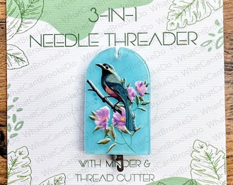 May include: A 3-in-1 needle threader with a light blue arched acrylic body featuring a printed image of a dark bird perched on a branch with pink flowers. The tool includes a metal minder and thread cutter, mounted on a white display card.