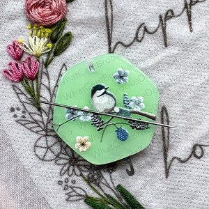 May include: A green plastic needle minder featuring a white bird and blue flowers on a branch. The needle minder is shaped like a hexagon and has the text "What Can't Brooke Do" written around the edge.