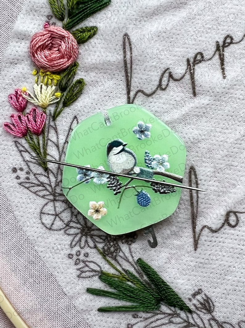 May include: A green plastic needle minder featuring a white bird and blue flowers on a branch. The needle minder is shaped like a hexagon and has the text "What Can't Brooke Do" written around the edge.