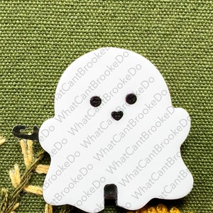 May include: White ghost-shaped ornament with black eyes and a black mouth. The ornament is on a green fabric background with yellow flowers.