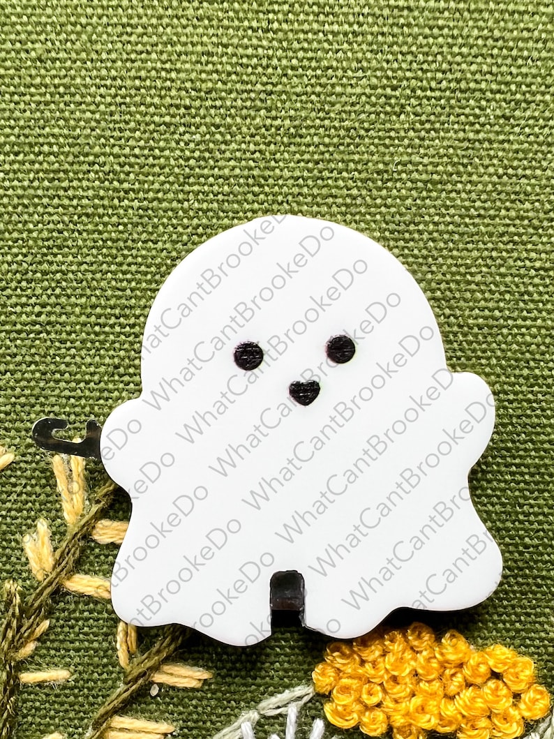 May include: White ghost-shaped ornament with black eyes and a black mouth. The ornament is on a green fabric background with yellow flowers.