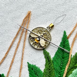 May include: A close-up of a gold bee charm with a yellow body and black wings, threaded on a needle with brown embroidery floss. The bee charm is surrounded by green embroidered leaves.