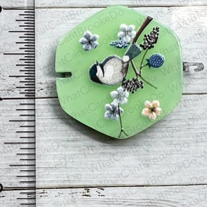 May include: A green resin cabochon featuring a painted image of a bluebird perched on a branch with white and pink flowers. The cabochon measures approximately 2.5 cm in diameter.