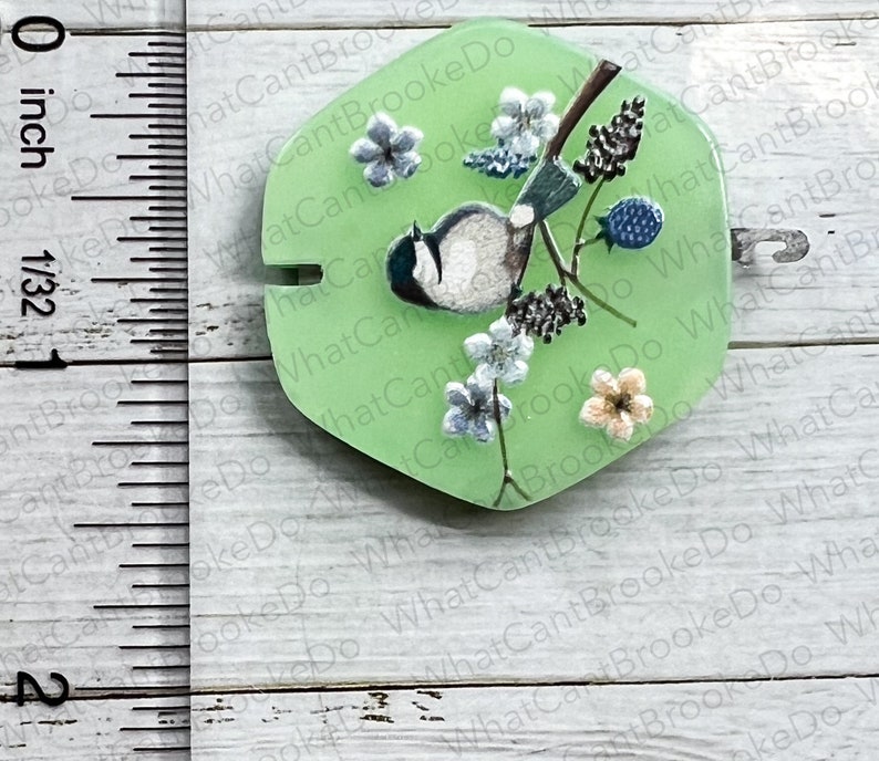 May include: A green resin cabochon featuring a painted image of a bluebird perched on a branch with white and pink flowers. The cabochon measures approximately 2.5 cm in diameter.