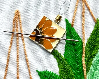 May include: Close-up of a needle threading through a small square-shaped embroidery piece with a floral design. The piece is attached to a gold-colored metal ring. The embroidery piece is surrounded by green and light green embroidery floss, creating a leafy design.