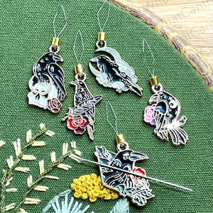 May include: A collection of six raven-themed stitch markers with gold-colored findings. Each marker features a different raven design, including a skull, roses, and a crescent moon. The markers are set against a green fabric background with embroidery.