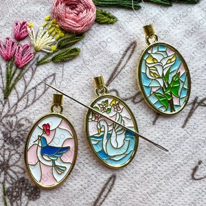 May include: Three oval-shaped, gold-toned, stained glass ornaments with different designs. The first ornament features a blue bird with a pink flower. The second ornament features a white swan on a blue lake. The third ornament features a white flower with green leaves.