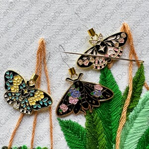 May include: Three gold-toned metal butterfly needle threaders with black, white, pink, and blue enamel details. The threaders are hanging from orange and white threads against a white fabric background.