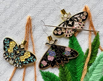 May include: Three gold-toned metal butterfly needle threaders with black, white, pink, and blue enamel details. The threaders are hanging from orange and white threads against a white fabric background.