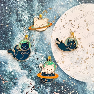 May include: Four enamel pin charms featuring cats in space. The charms are gold with blue and green enamel. One charm features a white cat in a spacesuit on a planet with a gold ring. Another charm features a black cat in a spacesuit floating in space. The other two charms feature white cats in spacesuits on planets with gold rings.