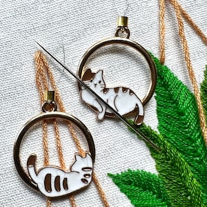 May include: Two gold embroidery hoops with white cats inside. The cats are outlined in gold and have brown stripes. The hoops are hanging from a white fabric background.