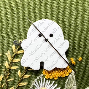May include: A white ghost-shaped embroidery hoop with a needle and thread going through it. The hoop is on a green fabric background with yellow and white embroidery.