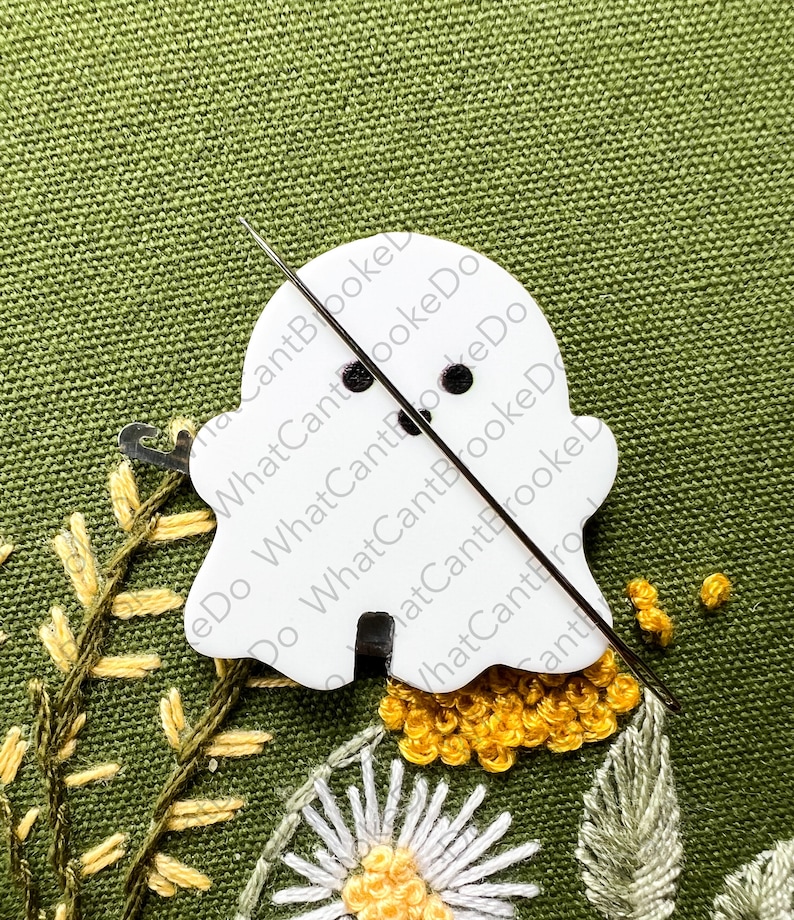 May include: A white ghost-shaped embroidery hoop with a needle and thread going through it. The hoop is on a green fabric background with yellow and white embroidery.