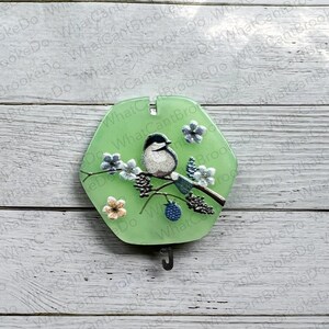 May include: A green hexagonal ornament featuring a bird and flowers on a branch. The ornament has a metal hook on the bottom.