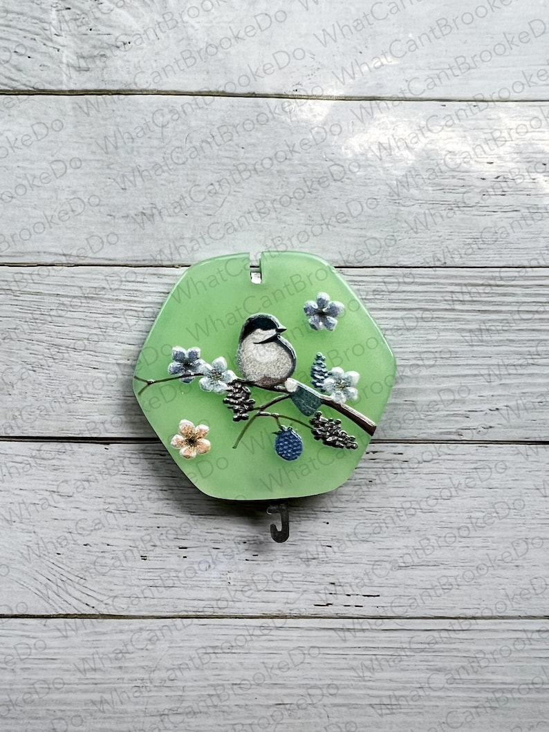 May include: A green hexagonal ornament featuring a bird and flowers on a branch. The ornament has a metal hook on the bottom.