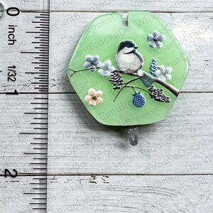 May include: A green hexagonal glass ornament featuring a painted scene of a bird perched on a branch with flowers and berries. The ornament has a silver hook for hanging.