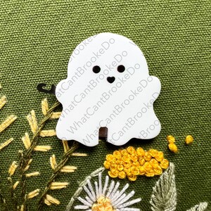 May include: White felt ghost with black eyes and a heart-shaped mouth. The ghost is on a green fabric background with yellow and white embroidered flowers.