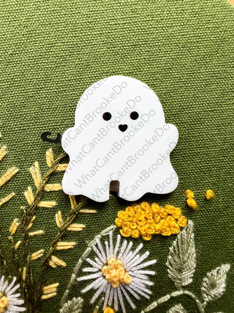 May include: White felt ghost with black eyes and a heart-shaped mouth. The ghost is on a green fabric background with yellow and white embroidered flowers.