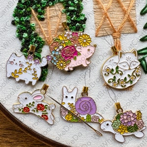 May include: Six colorful needle threaders with different animal and nature designs. The threaders are made of metal and have a gold finish. The designs include a cat, a hedgehog, a fox, a rabbit, a snail, and a turtle.