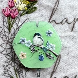 May include: A green hexagonal ornament featuring a white bird perched on a branch with blue flowers and black berries. The ornament has a metal hook and the text "ReDo What Can BrookeDo What CantBrookeDo" is printed around the edge.