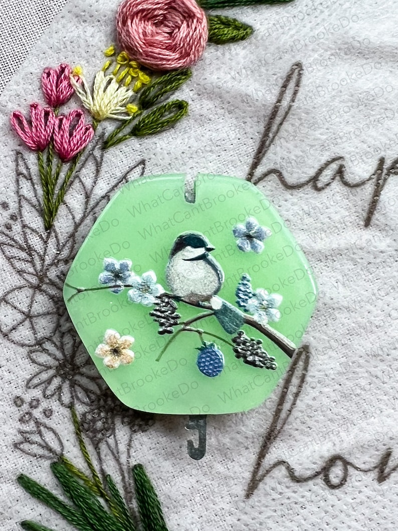 May include: A green hexagonal ornament featuring a white bird perched on a branch with blue flowers and black berries. The ornament has a metal hook and the text "ReDo What Can BrookeDo What CantBrookeDo" is printed around the edge.