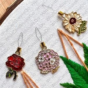 May include: Three gold-toned needle threaders with floral designs. The threaders feature a red rose, a pink hydrangea, and a yellow sunflower. Each design has a gold-toned loop at the top.