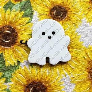 May include: White ghost-shaped button with black eyes and a small mouth. The button is on a fabric background with a sunflower pattern.