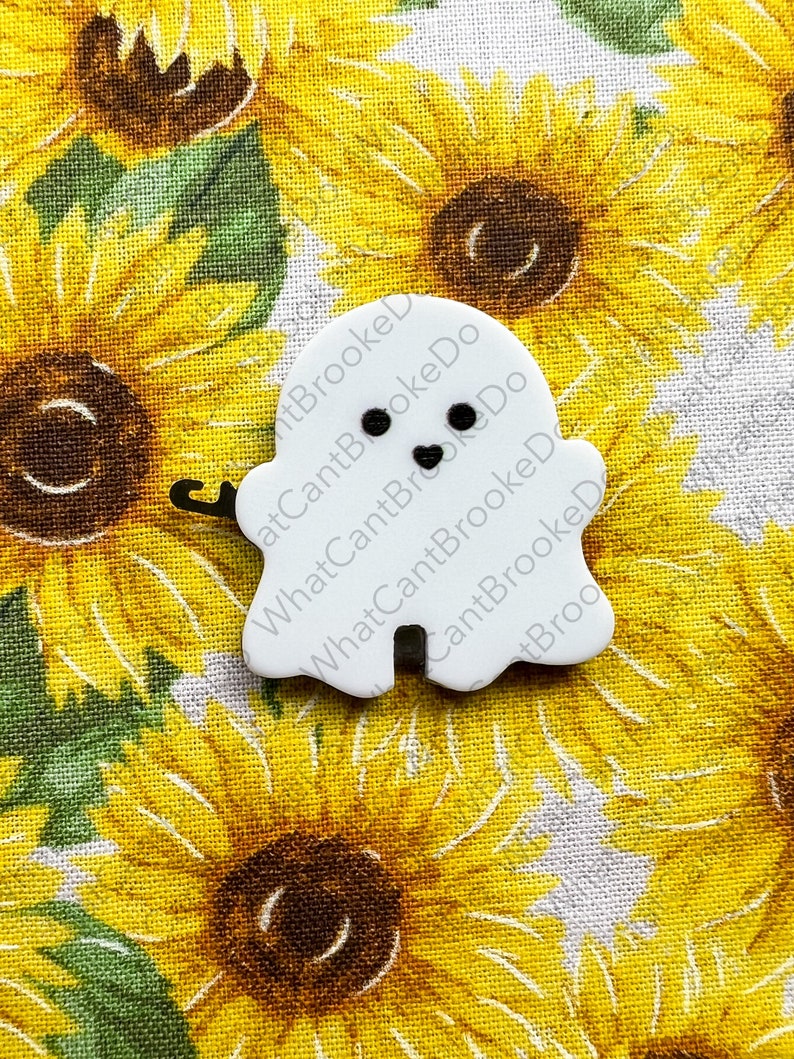 May include: White ghost-shaped button with black eyes and a small mouth. The button is on a fabric background with a sunflower pattern.