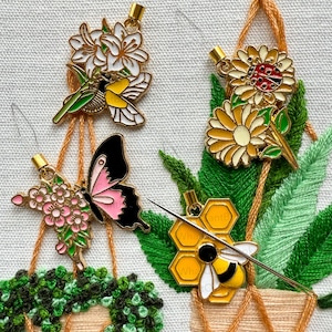 May include: Four embroidered floral designs with a bee, butterfly, hummingbird, and ladybug. Each design is attached to a piece of embroidery floss and is being stitched onto a piece of fabric.