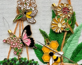 May include: Four embroidered floral designs with a bee, butterfly, hummingbird, and ladybug. Each design is attached to a piece of embroidery floss and is being stitched onto a piece of fabric.
