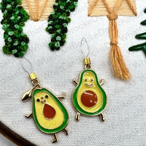 May include: Two gold-colored avocado charms with green and yellow enamel, and a brown seed. The charms have small arms and legs. The background is a white fabric with embroidery of green leaves and a tan square.
