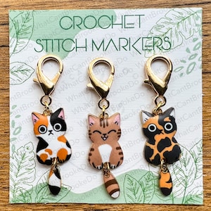 May include: Three gold crochet stitch markers with cat charms. The cats are a calico, a tabby, and a tortoiseshell. Each cat has a gold tail.