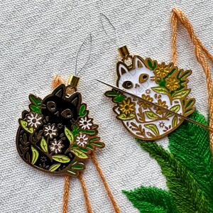 May include: Two gold-colored cat needle minders with black and white cats surrounded by green leaves and white flowers. The cats are holding a needle in their paws.