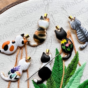 May include: Six needle threaders shaped like cats with different patterns and colors. The cats are on a white fabric background with green leaves.