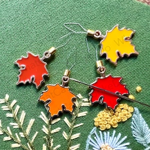May include: Four gold-colored metal maple leaf charms with red, orange, and yellow enamel. The charms are attached to thin metal wires with gold beads.